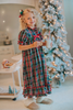 A young girl is wearing The Original Clara Nutcracker Gown in black plaid featuring black plaid ruffles and a red bow. The plaid has white, red and green with a black background. The buttery soft fabric is cozy and sensory friendly.  The puff sleeves with ribbon detail and the ruffles at the hem add a traditional classic look. 