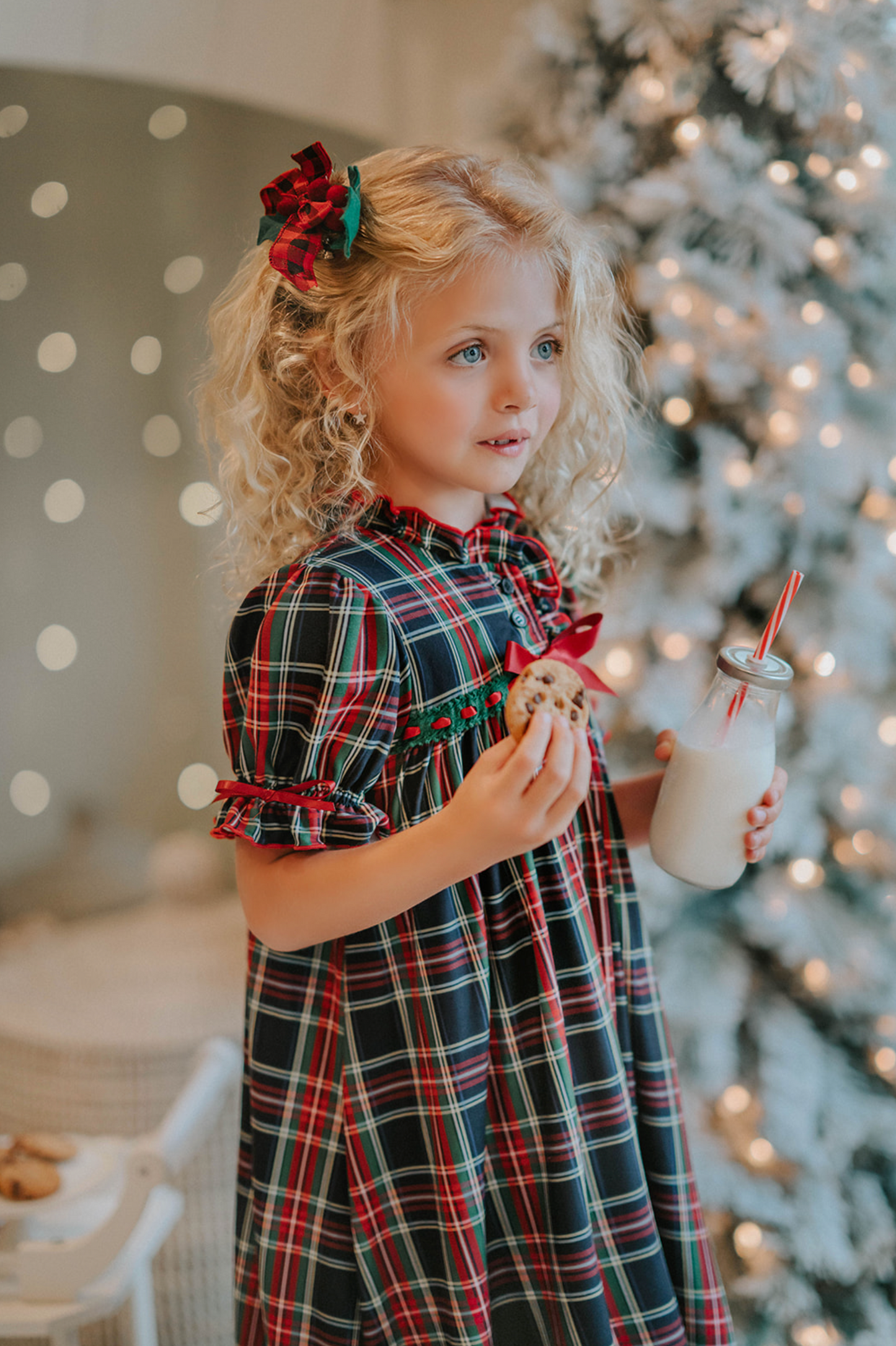 A young girl is wearing The Original Clara Nutcracker Gown in black plaid featuring black plaid ruffles and a red bow. The plaid has white, red and green with a black background. The buttery soft fabric is cozy and sensory friendly.  The puff sleeves with ribbon detail and the ruffles at the hem add a traditional classic look. 