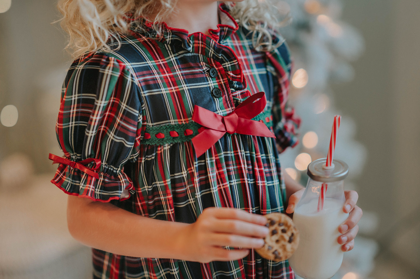 A young girl is wearing The Original Clara Nutcracker Gown in black plaid featuring black plaid ruffles and a red bow. The plaid has white, red and green with a black background. The buttery soft fabric is cozy and sensory friendly.  The puff sleeves with ribbon detail and the ruffles at the hem add a traditional classic look. 