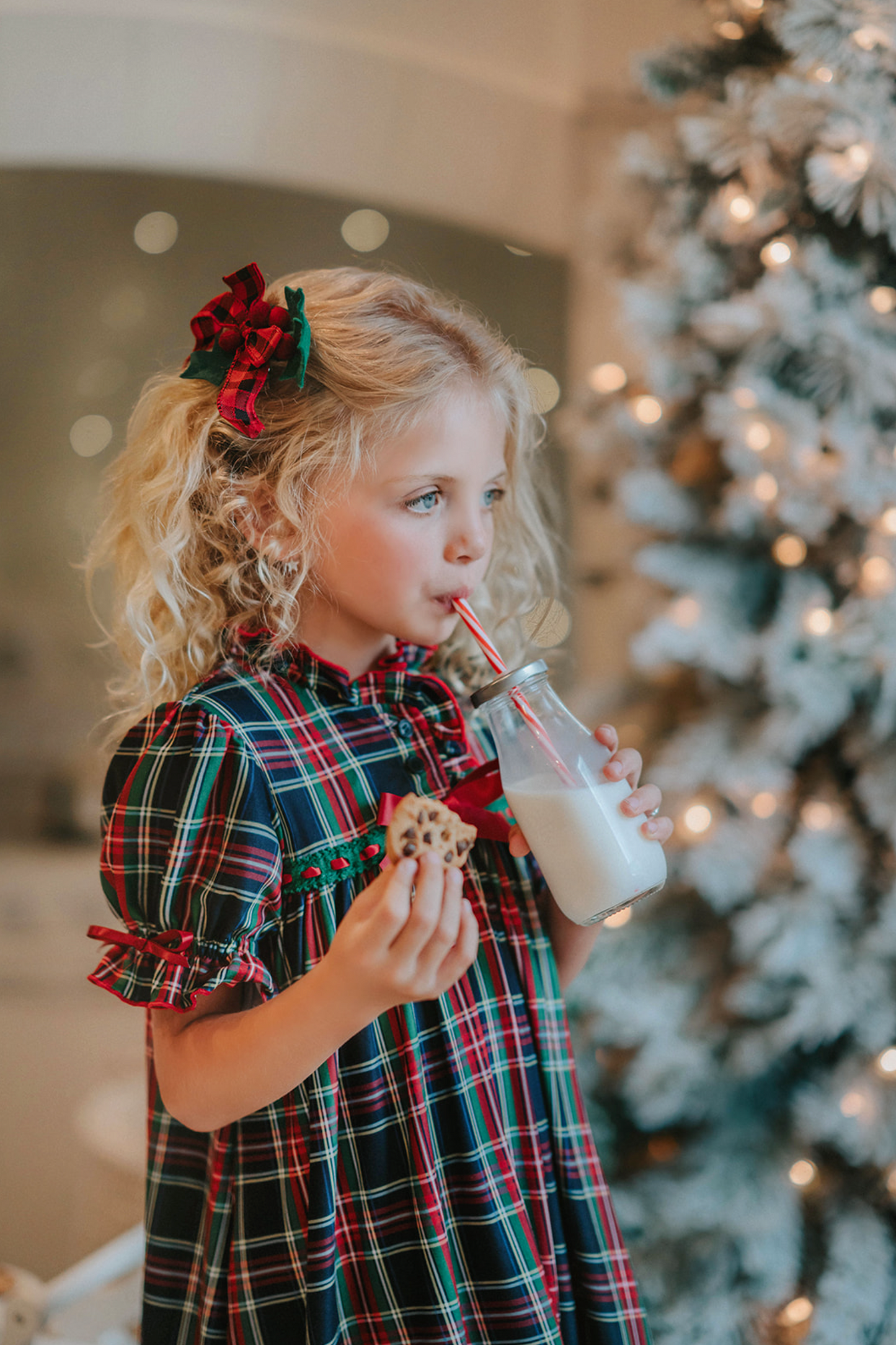 A young girl is wearing The Original Clara Nutcracker Gown in black plaid featuring black plaid ruffles and a red bow. The plaid has white, red and green with a black background. The buttery soft fabric is cozy and sensory friendly.  The puff sleeves with ribbon detail and the ruffles at the hem add a traditional classic look. 