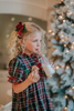 A young girl is wearing The Original Clara Nutcracker Gown in black plaid featuring black plaid ruffles and a red bow. The plaid has white, red and green with a black background. The buttery soft fabric is cozy and sensory friendly.  The puff sleeves with ribbon detail and the ruffles at the hem add a traditional classic look. 