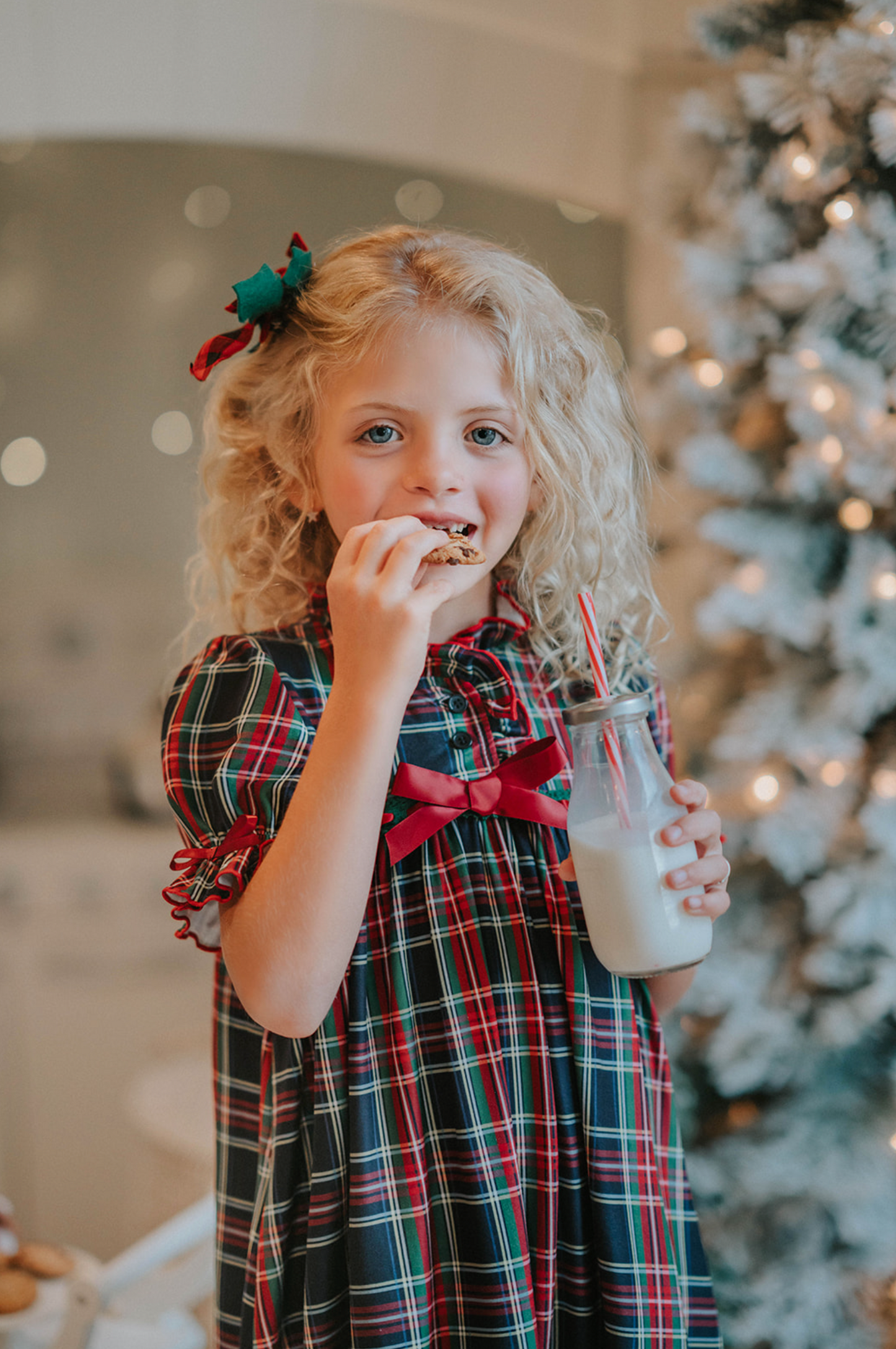 A young girl is wearing The Original Clara Nutcracker Gown in black plaid featuring black plaid ruffles and a red bow. The plaid has white, red and green with a black background. The buttery soft fabric is cozy and sensory friendly.  The puff sleeves with ribbon detail and the ruffles at the hem add a traditional classic look. 