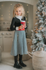 A young girl with curly blonde hair stands holding a red Christmas ornament. She is wearing a black top, a red belt, and the White Christmas Houndstooth Dress that adds a festive touch to the scene, with a decorated Christmas tree in the background. She appears thoughtful in the cozy, warmly lit room—a truly timeless photo.