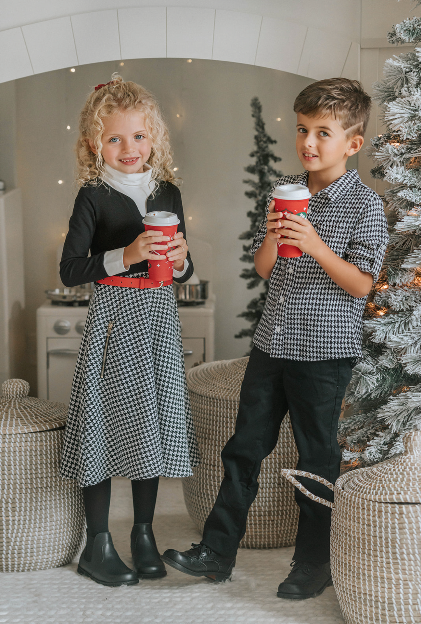 A young girl with curly blonde hair stands holding a red Christmas ornament. She is wearing a black top, a red belt, and the White Christmas Houndstooth Dress that adds a festive touch to the scene, with a decorated Christmas tree in the background. She appears thoughtful in the cozy, warmly lit room—a truly timeless photo.