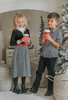 A young boy stands in a festive setting with a decorated Christmas tree and twinkling lights in the background. He is wearing a White Christmas Houndstooth Shirt and black pants, with one hand in his pocket, smiling at the camera, as if waiting for Santa to arrive.