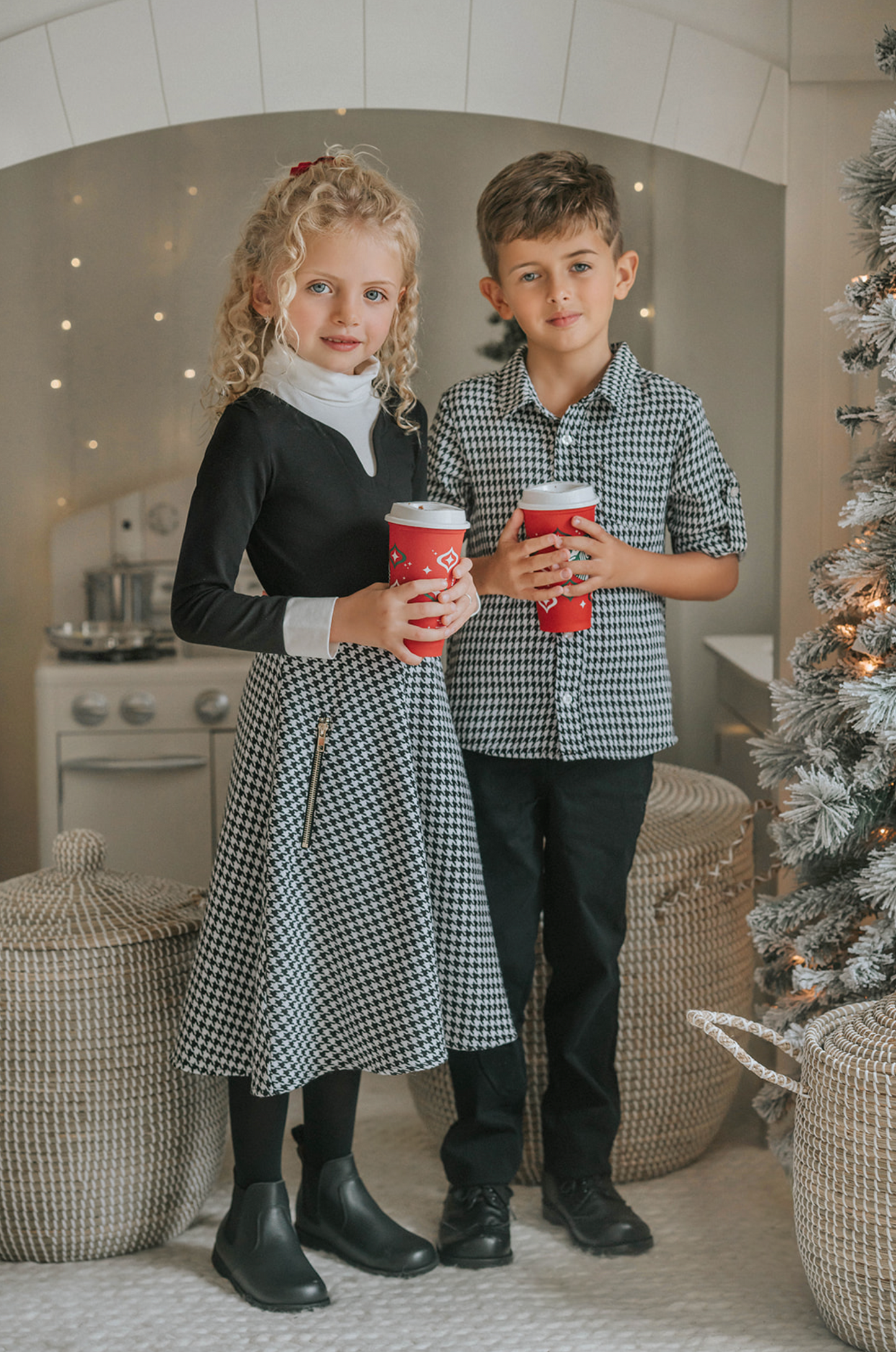 A young boy stands in a festive setting with a decorated Christmas tree and twinkling lights in the background. He is wearing a White Christmas Houndstooth Shirt and black pants, with one hand in his pocket, smiling at the camera, as if waiting for Santa to arrive.