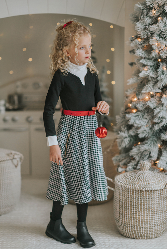 A young girl with curly blonde hair stands holding a red Christmas ornament. She is wearing a black top, a red belt, and the White Christmas Houndstooth Dress that adds a festive touch to the scene, with a decorated Christmas tree in the background. She appears thoughtful in the cozy, warmly lit room—a truly timeless photo.