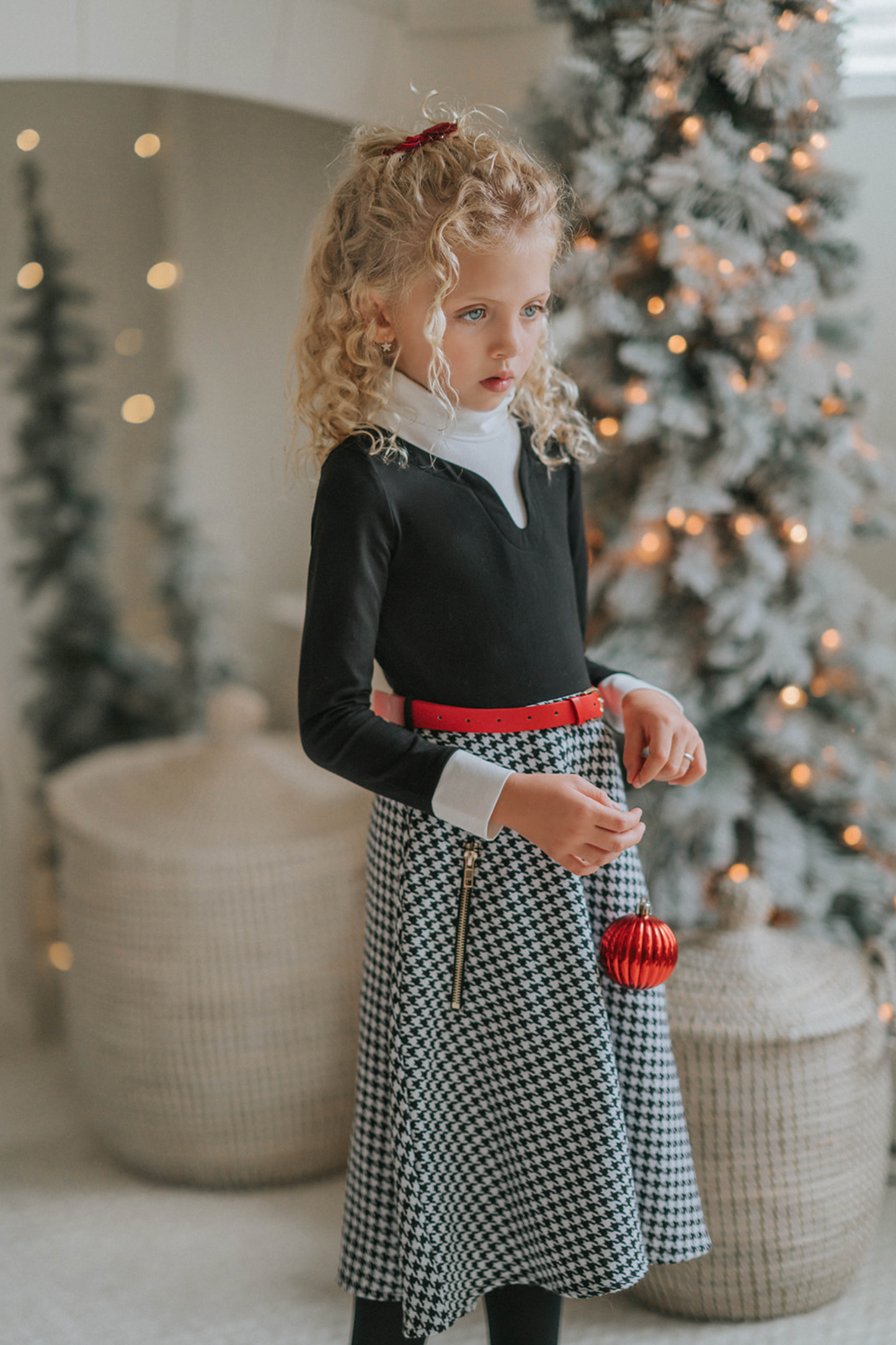 A young girl with curly blonde hair stands holding a red Christmas ornament. She is wearing a black top, a red belt, and the White Christmas Houndstooth Dress that adds a festive touch to the scene, with a decorated Christmas tree in the background. She appears thoughtful in the cozy, warmly lit room—a truly timeless photo.