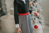 A young girl with curly blonde hair stands holding a red Christmas ornament. She is wearing a black top, a red belt, and the White Christmas Houndstooth Dress that adds a festive touch to the scene, with a decorated Christmas tree in the background. She appears thoughtful in the cozy, warmly lit room—a truly timeless photo.