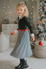 A young girl with curly blonde hair stands holding a red Christmas ornament. She is wearing a black top, a red belt, and the White Christmas Houndstooth Dress that adds a festive touch to the scene, with a decorated Christmas tree in the background. She appears thoughtful in the cozy, warmly lit room—a truly timeless photo.