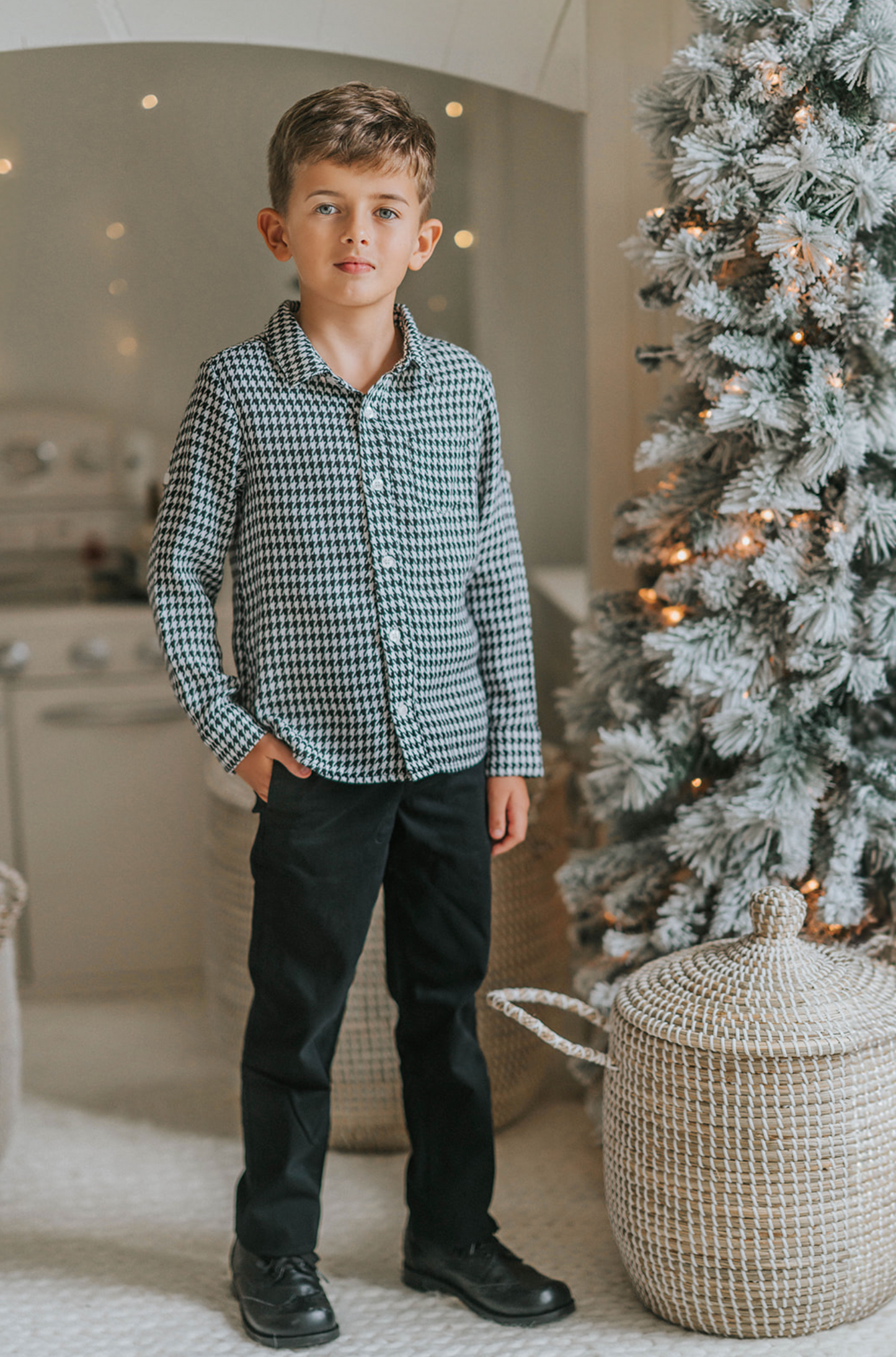 A young boy stands in a festive setting with a decorated Christmas tree and twinkling lights in the background. He is wearing a White Christmas Houndstooth Shirt and black pants, with one hand in his pocket, smiling at the camera, as if waiting for Santa to arrive.