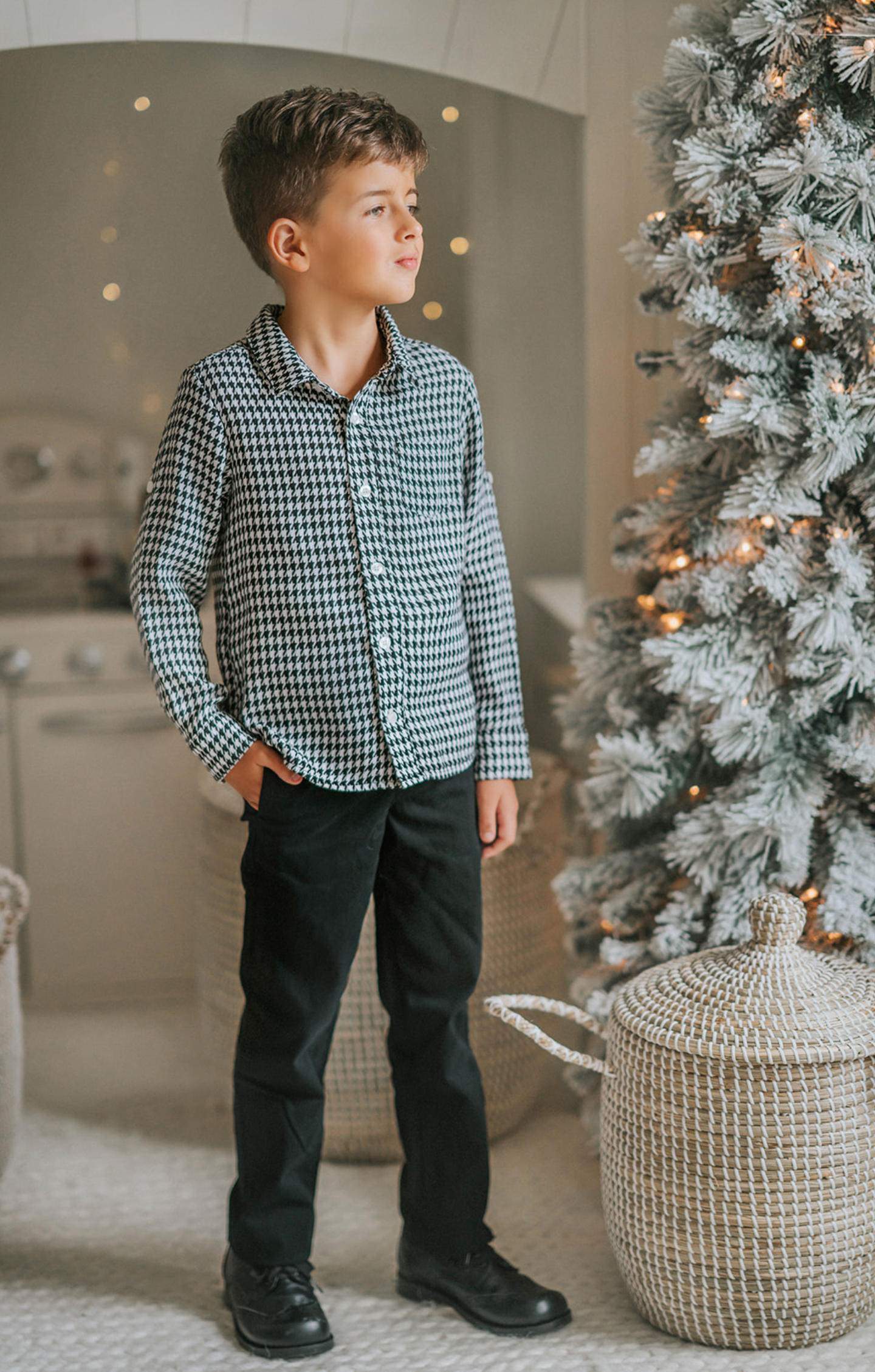 A young boy stands in a festive setting with a decorated Christmas tree and twinkling lights in the background. He is wearing a White Christmas Houndstooth Shirt and black pants, with one hand in his pocket, smiling at the camera, as if waiting for Santa to arrive.