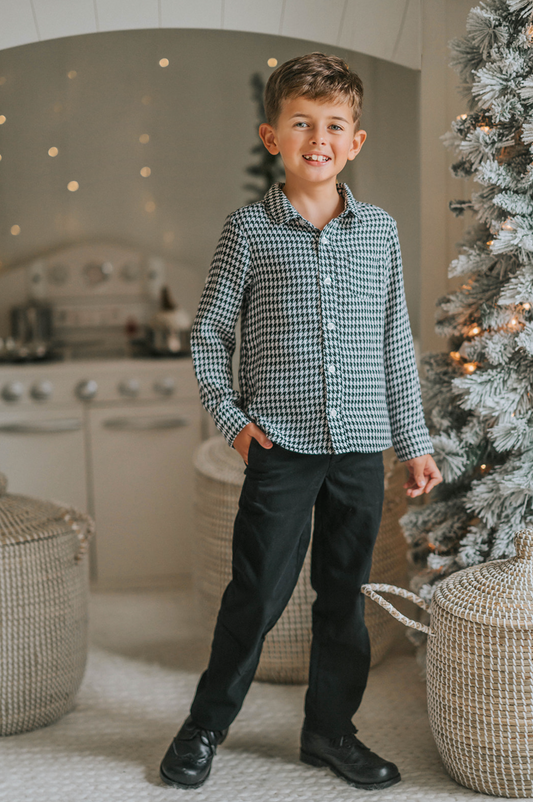 A young boy stands in a festive setting wearing a White Christmas Houndstooth Shirt and black pants. The long sleeve oxford button up shirt has a small back and white houndstooth print and matches a dress for a matching sibling set. 