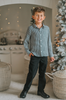 A young boy stands in a festive setting wearing a White Christmas Houndstooth Shirt and black pants. The long sleeve oxford button up shirt has a small back and white houndstooth print and matches a dress for a matching sibling set. 