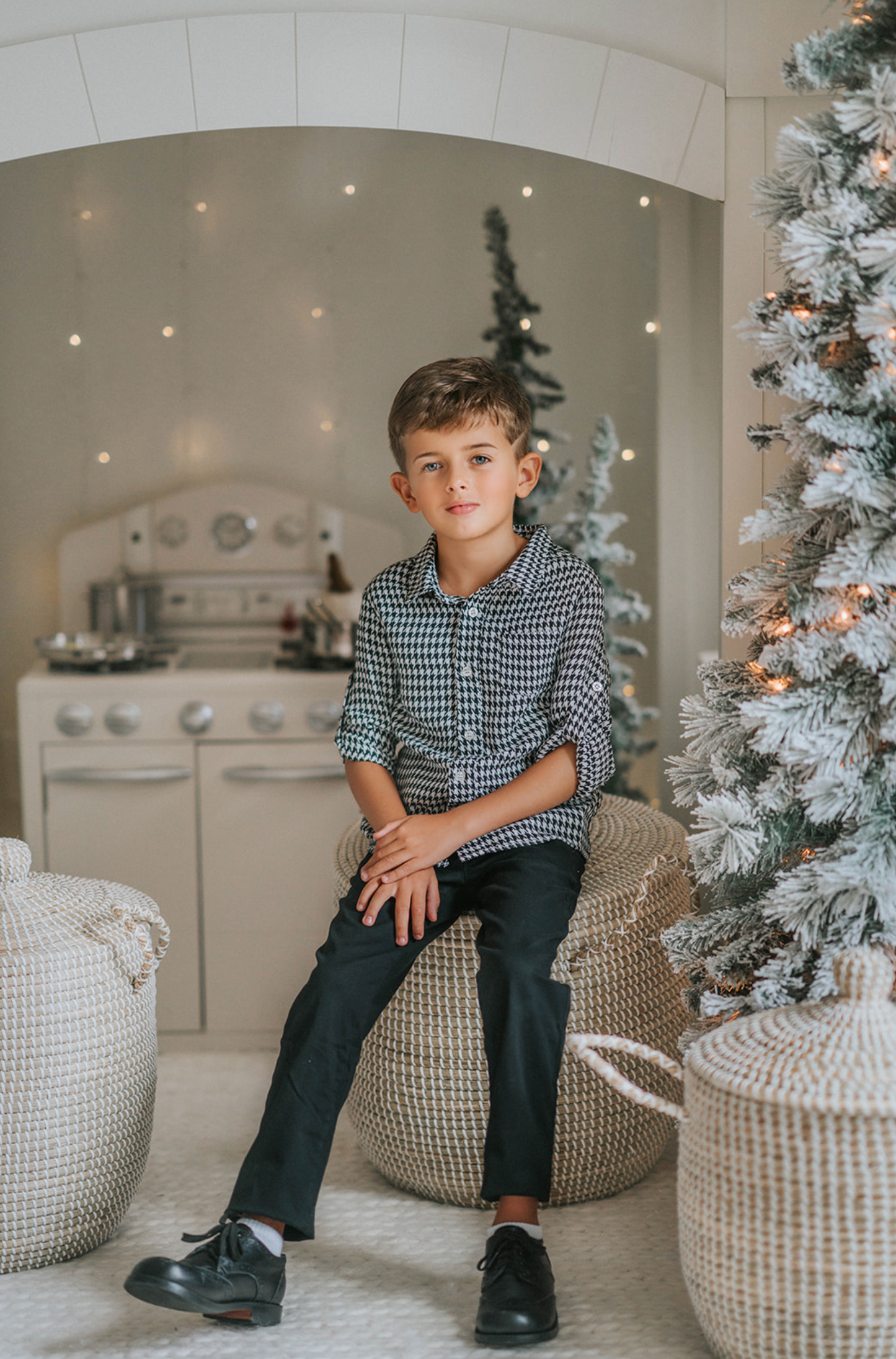 A young boy stands in a festive setting with a decorated Christmas tree and twinkling lights in the background. He is wearing a White Christmas Houndstooth Shirt and black pants, with one hand in his pocket, smiling at the camera, as if waiting for Santa to arrive.