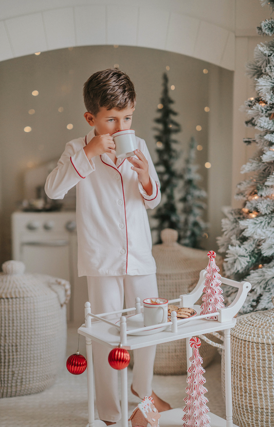 A young boy is wearing a traditional and classic 2pc lounge set in Ivory Bisque.  The top has maroon piping.  The buttery soft fabric and sibling matching option make this a perfect holiday set. 