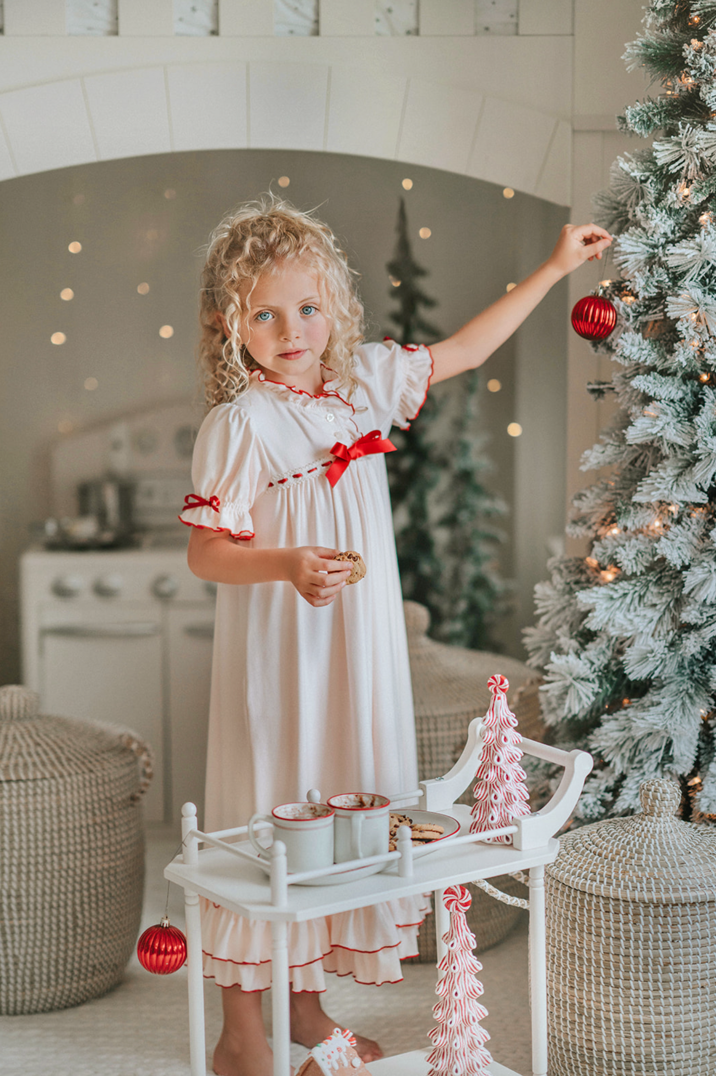 A young girl is wearing The Original Clara Nutcracker Gown in Ivory Bisque, featuring ivory bisque ruffles and a maroon bow. The buttery soft fabric is cozy and sensory friendly.  The puff sleeves with ribbon detail and the ruffles at the hem add a traditional classic look. 