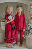 A young boy in buttery soft Unisex Pj's in Merry Maroon holds a nutcracker doll.  The traditional 2 piece pajama set has ivory piping and maroon buttons down the front. A girl in matching pajamas with curly hair is partially visible in the background.