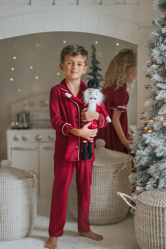 A young boy in buttery soft Unisex Pj's in Merry Maroon holds a nutcracker doll, standing near a decorated Christmas tree and a white wicker basket. A girl in matching pajamas with curly hair is partially visible in the background, reflecting festive holiday decor with blurred lights from the Christmas Lounge Collection.