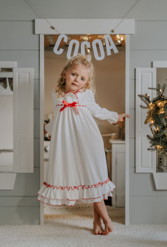 A young child is wearing The Original Clara Nutcracker Christmas Gown Long Sleeve in White. The front has two buttons at the collar with red trim detail. There is a bow and ribbon along the chest. The bottom has two ruffles with red trim detail. The fabric is a buttery soft polyester. 