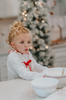 A young child with light curly hair stands indoors wearing The Original Clara Nutcracker Christmas Gown Long Sleeve in White from the Christmas Lounge Collection. Behind them, a white doorway decorated with a sign reading "Cocoa" is visible. There is also a small Christmas tree adorned with lights in the background, adding to the festive atmosphere.