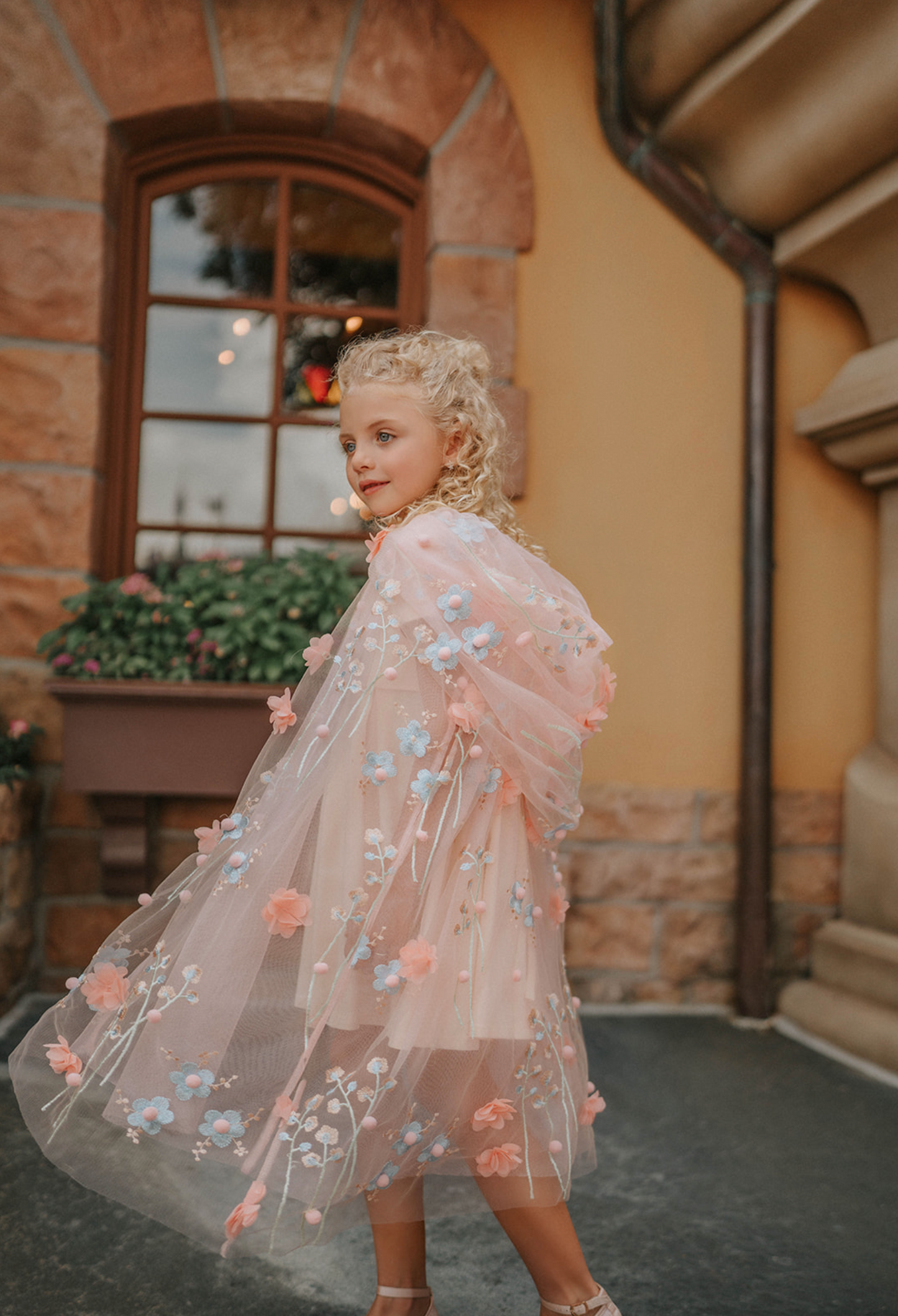 Young girl wearing a very soft pink tulle cape with hood.  There are beautiful floral flowers with pink petals and light blue flowers with pink fuzzy centers.  Greenery in light green is scattered around the flowers. 