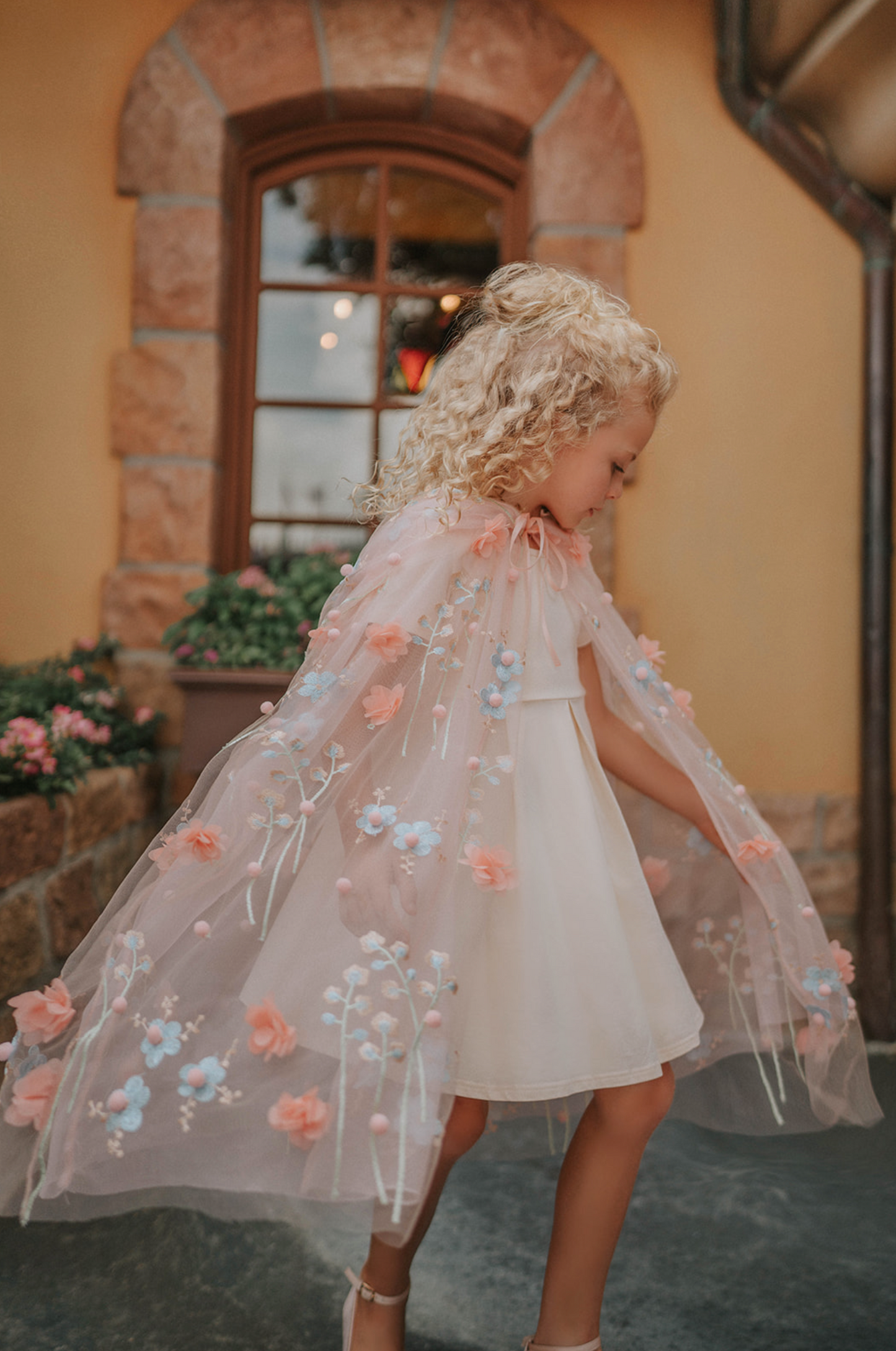 Young girl wearing a very soft pink tulle cape with hood.  There are beautiful floral flowers with pink petals and light blue flowers with pink fuzzy centers.  Greenery in light green is scattered around the flowers. 