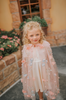 Young girl wearing a very soft pink tulle cape with hood.  There are beautiful floral flowers with pink petals and light blue flowers with pink fuzzy centers.  Greenery in light green is scattered around the flowers. 