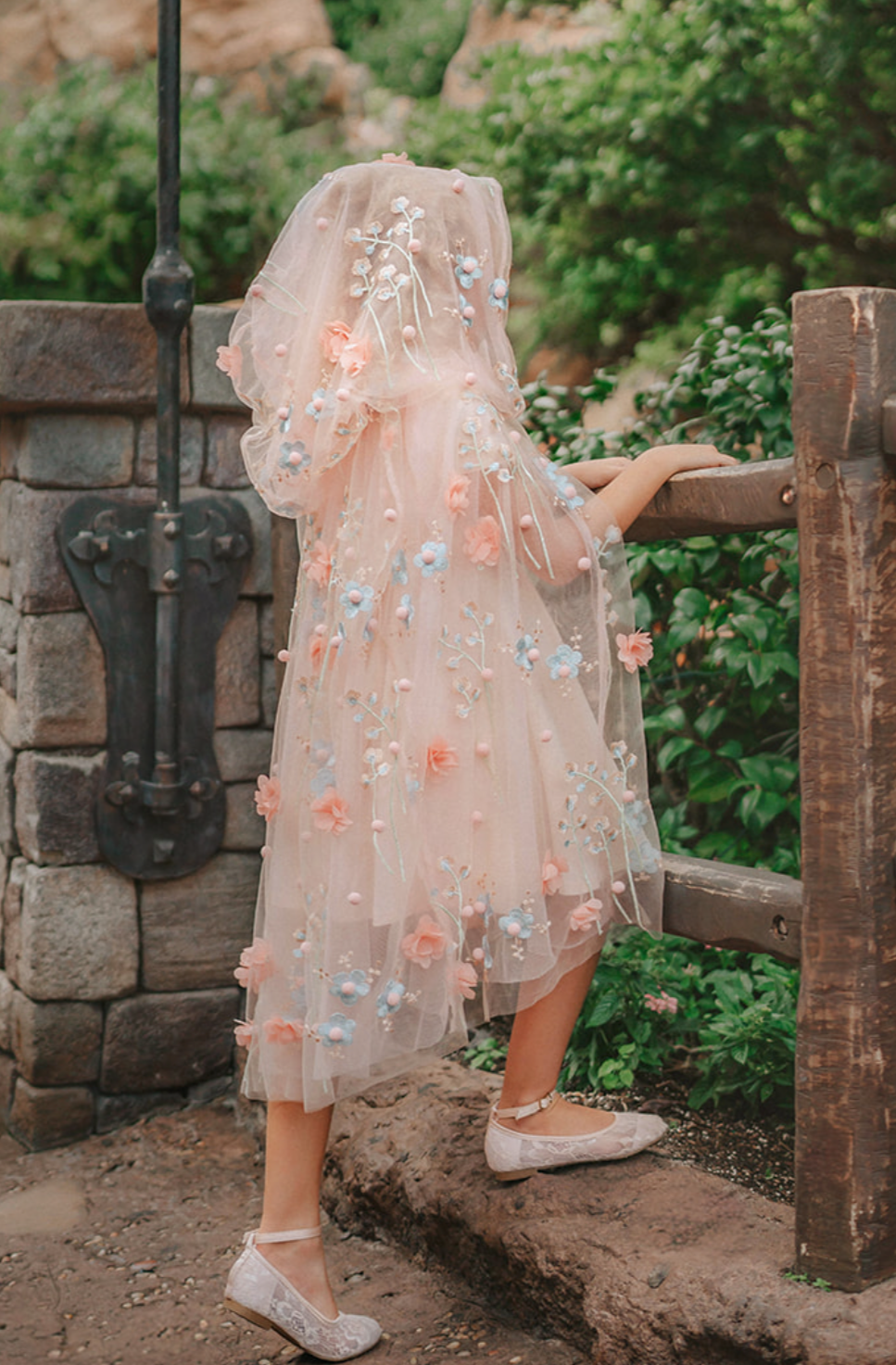 Young girl wearing a very soft pink tulle cape with hood.  There are beautiful floral flowers with pink petals and light blue flowers with pink fuzzy centers.  Greenery in light green is scattered around the flowers. 