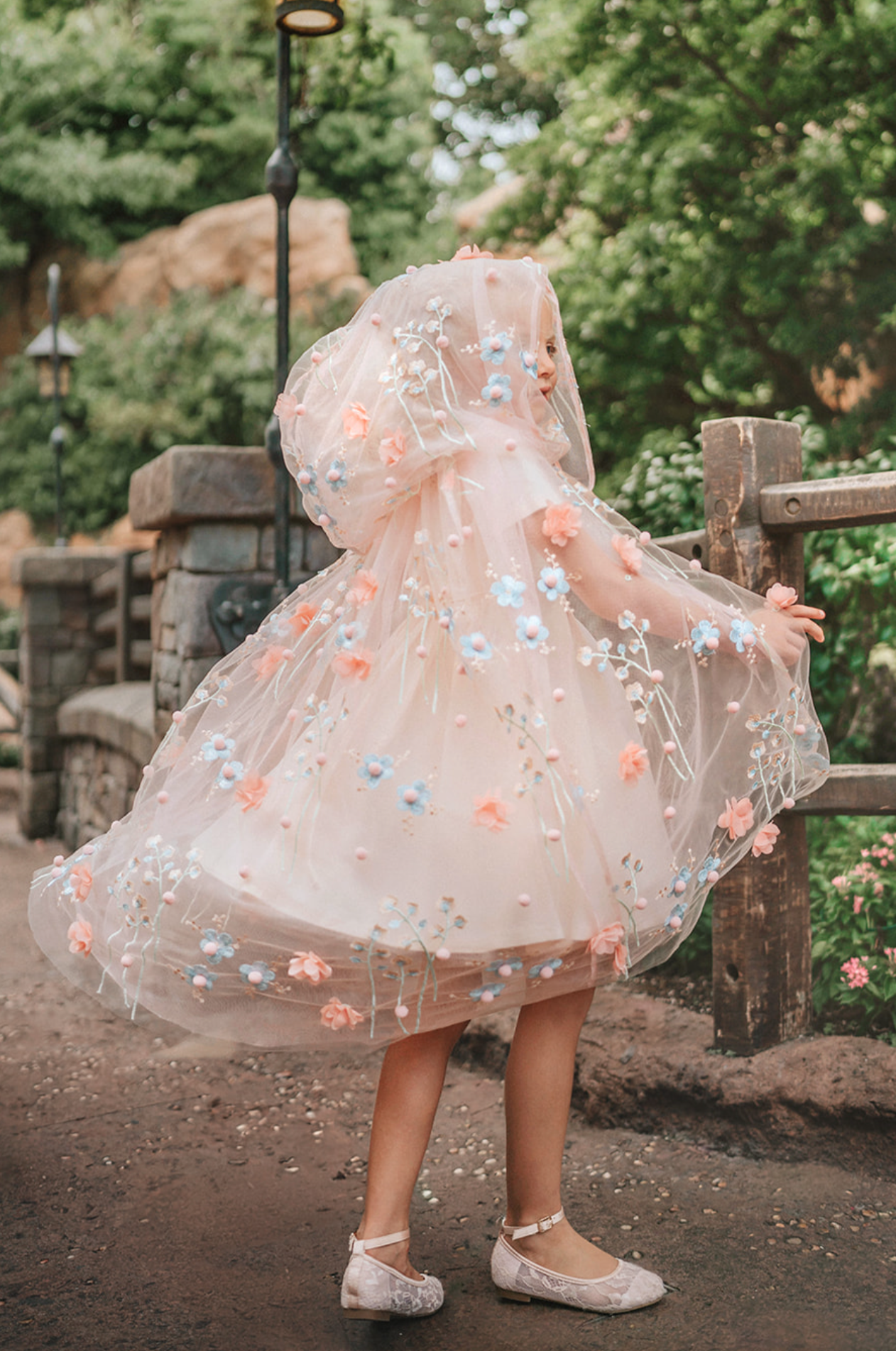 Young girl wearing a very soft pink tulle cape with hood.  There are beautiful floral flowers with pink petals and light blue flowers with pink fuzzy centers.  Greenery in light green is scattered around the flowers. 