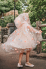 Young girl wearing a very soft pink tulle cape with hood.  There are beautiful floral flowers with pink petals and light blue flowers with pink fuzzy centers.  Greenery in light green is scattered around the flowers. 