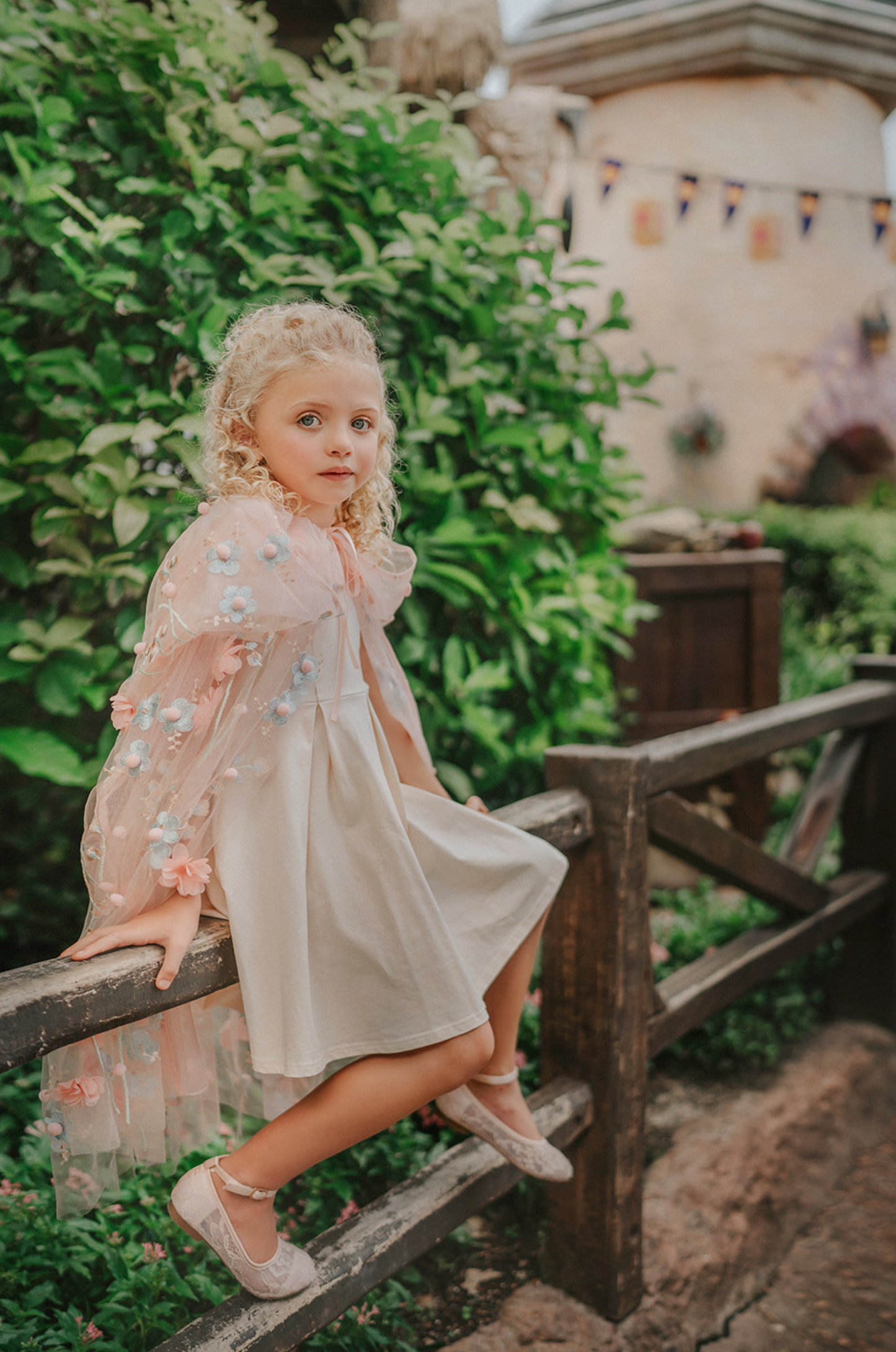 Young girl wearing a very soft pink tulle cape with hood.  There are beautiful floral flowers with pink petals and light blue flowers with pink fuzzy centers.  Greenery in light green is scattered around the flowers. 