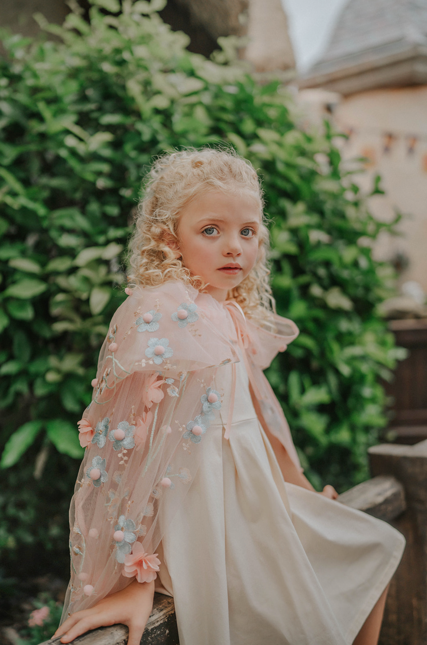 Young girl wearing a very soft pink tulle cape with hood.  There are beautiful floral flowers with pink petals and light blue flowers with pink fuzzy centers.  Greenery in light green is scattered around the flowers. 