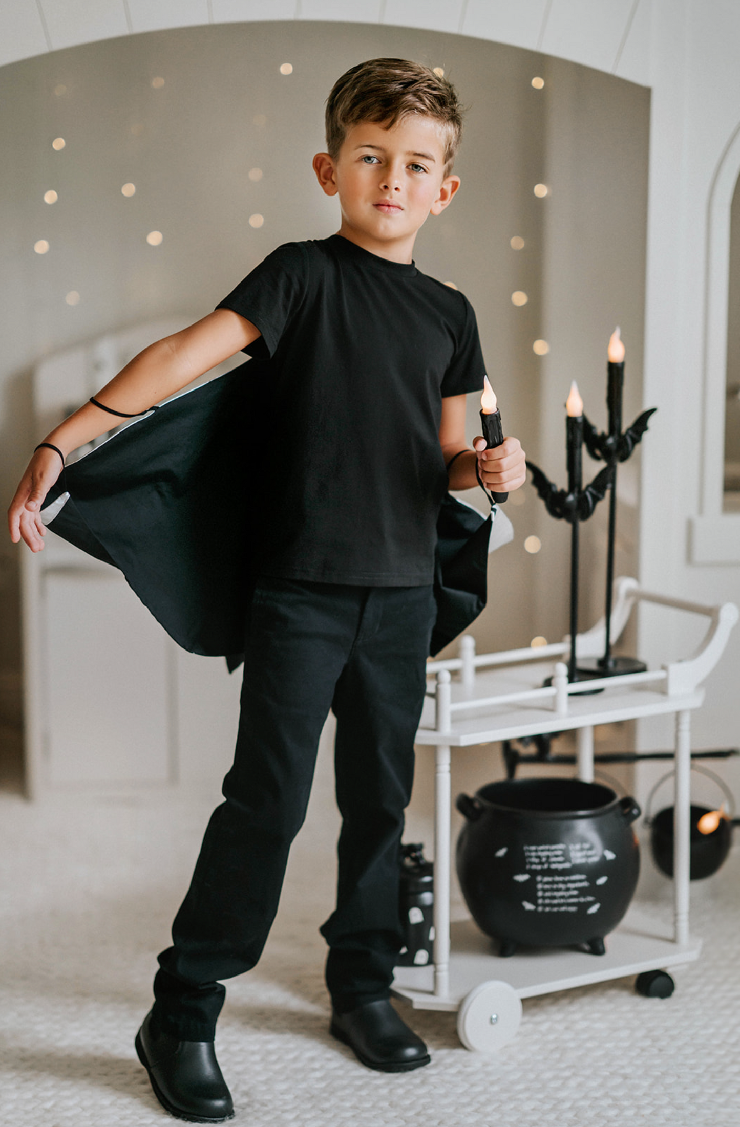 A young boy dressed in a Boys Bat Wing Shirt stands in a room decorated for Halloween.