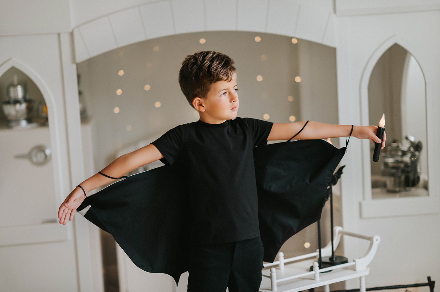 A young boy with short brown hair is dressed in a black Boys Bat Wing Shirt, extending his arms to reveal the bat-like cape. The removable cape makes this an ideal Halloween costume from our unisex collection.