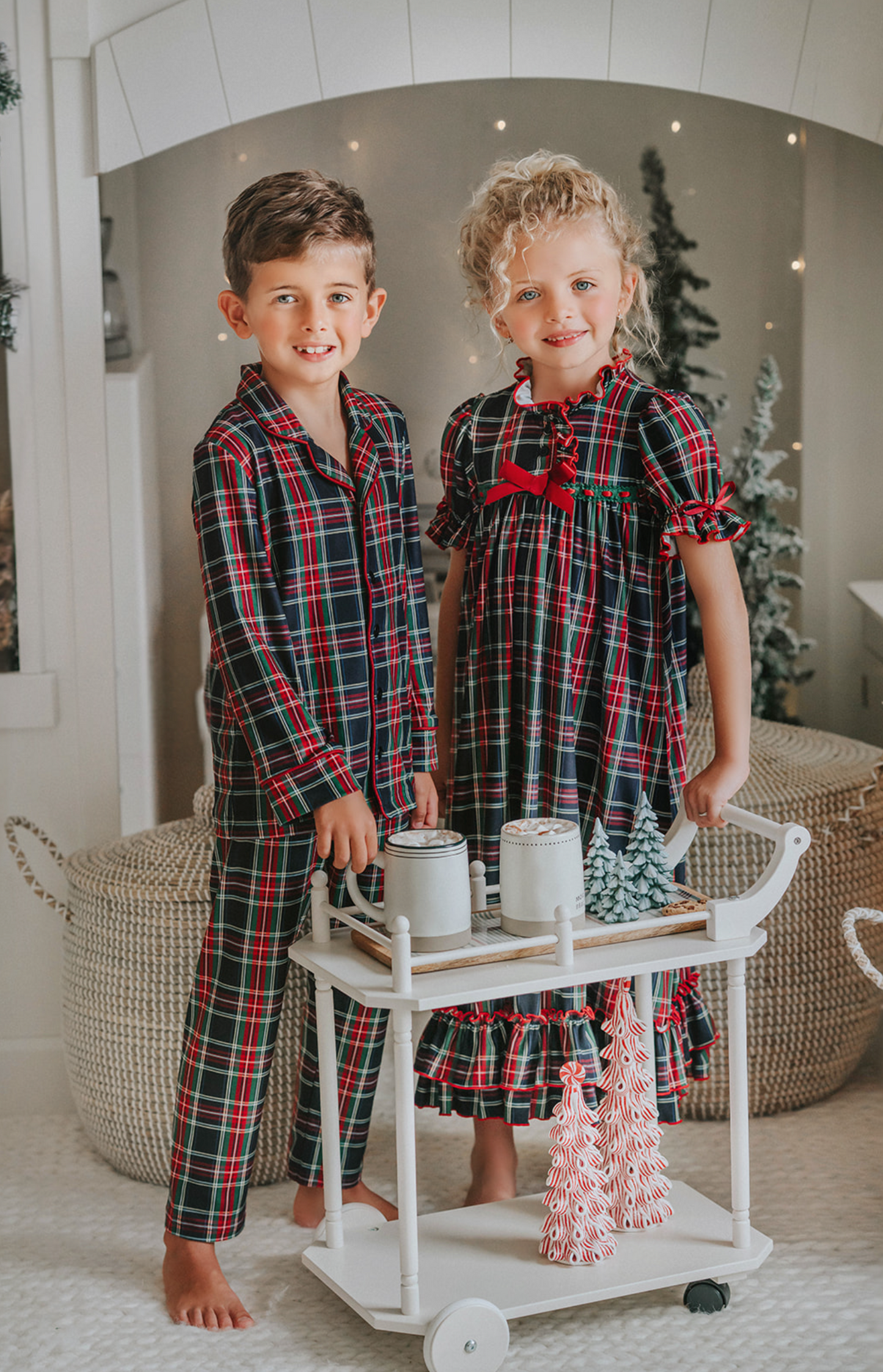 A young boy in Unisex Pj's in Black Plaid stands beside a small white cart with miniature Christmas trees on top. The background features a festive, cozy room with twinkling lights, a decorated Christmas tree, and holiday-themed decor.