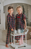 A young boy in Unisex Pj's in Black Plaid stands beside a small white cart with miniature Christmas trees on top. The background features a festive, cozy room with twinkling lights, a decorated Christmas tree, and holiday-themed decor.