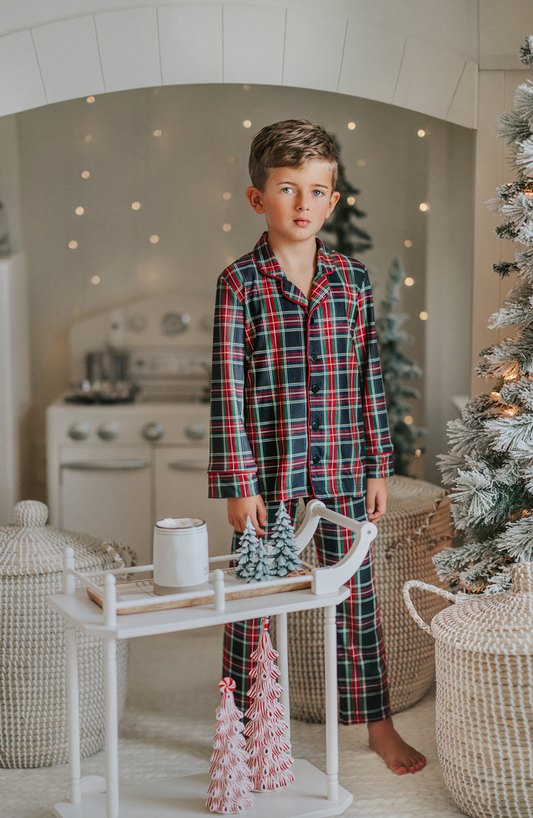 A young boy in Unisex Pj's in Black Plaid stands beside a small white cart with miniature Christmas trees on top. The background features a festive, cozy room with twinkling lights, a decorated Christmas tree, and holiday-themed decor.