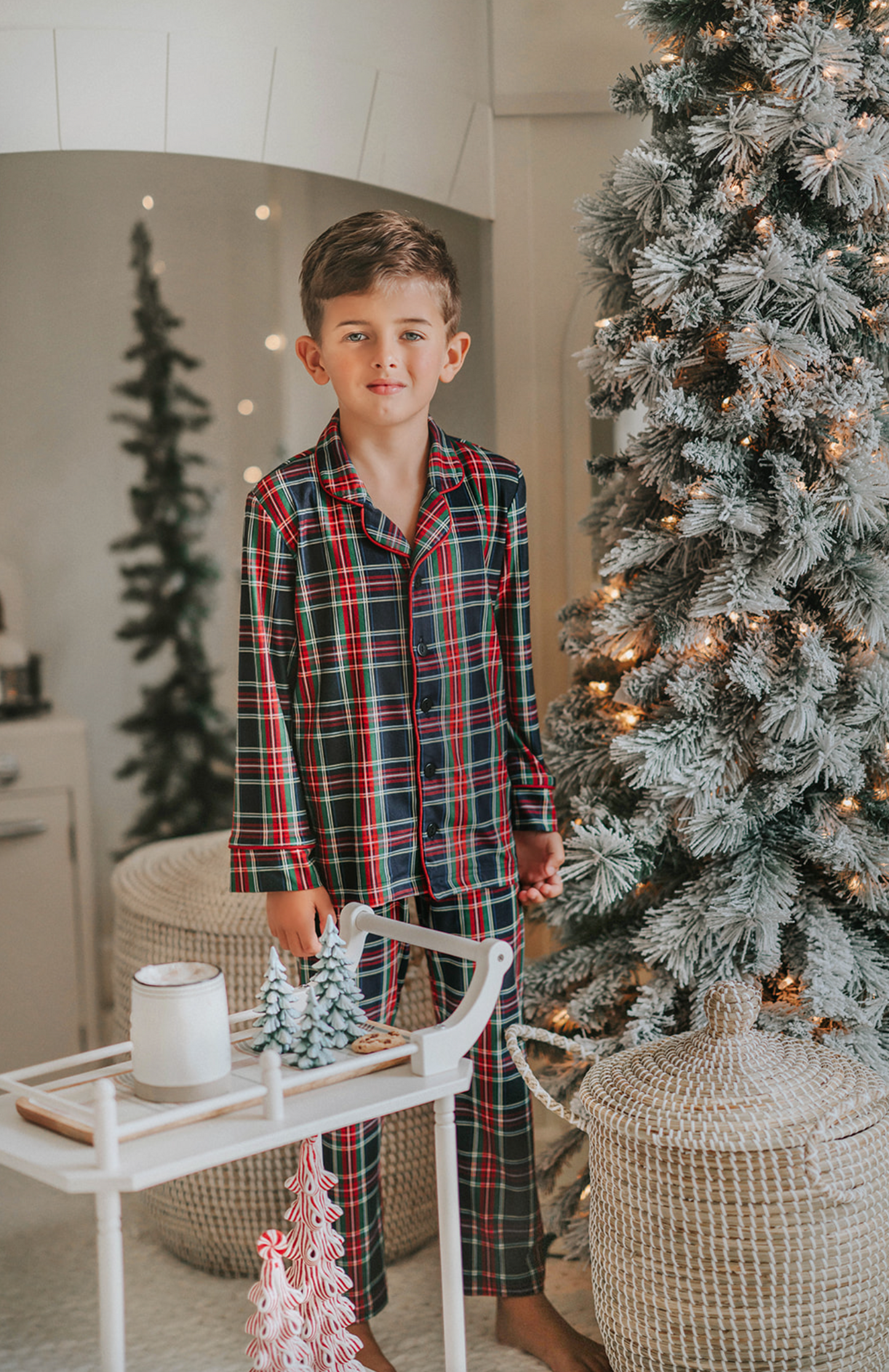 A young boy in Unisex Pj's in Black Plaid stands beside a small white cart with miniature Christmas trees on top. The background features a festive, cozy room with twinkling lights, a decorated Christmas tree, and holiday-themed decor.