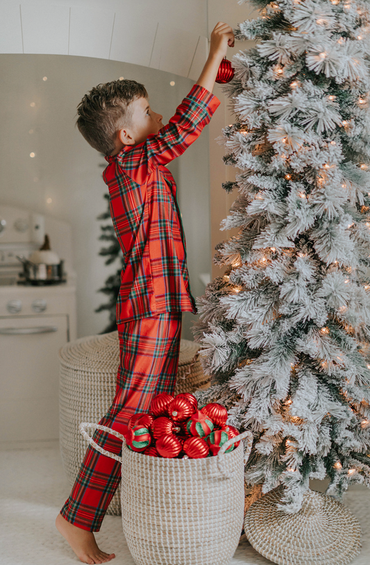 A young child in Unisex Pj's in Red Plaid decorates a tall, frosted Christmas tree with red ornaments. A basket filled with more red ornaments is placed nearby on the floor. The setting is a cozy, softly lit room, perfect for capturing heartwarming Christmas photos.