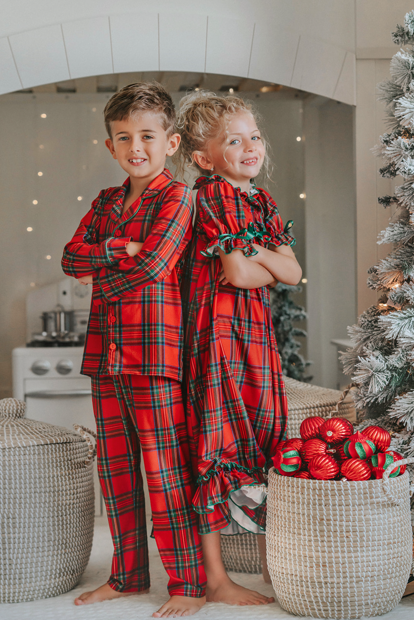 A young child in Unisex Pj's in Red Plaid decorates a Christmas tree. The plaid has green, white and black over a bright red.  The classic 2 piece pajamas have green piping and red buttons down the front of the shirt. 