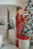 A young child in Unisex Pj's in Red Plaid decorates a Christmas tree. The plaid has green, white and black over a bright red.  The classic 2 piece pajamas have green piping and red buttons down the front of the shirt. 