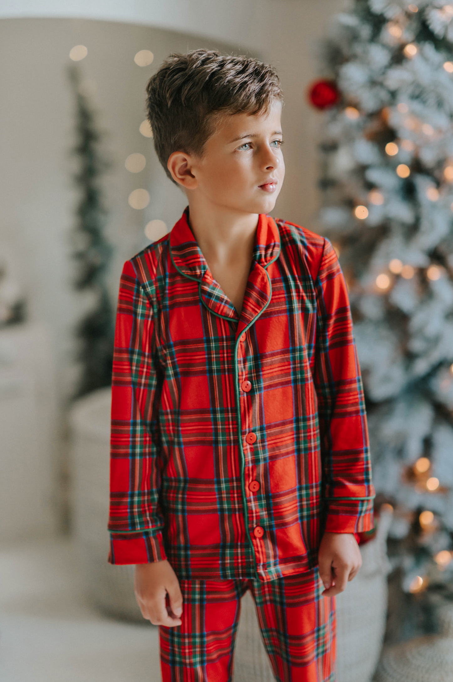 A young child in Unisex Pj's in Red Plaid decorates a Christmas tree. The plaid has green, white and black over a bright red.  The classic 2 piece pajamas have green piping and red buttons down the front of the shirt. 