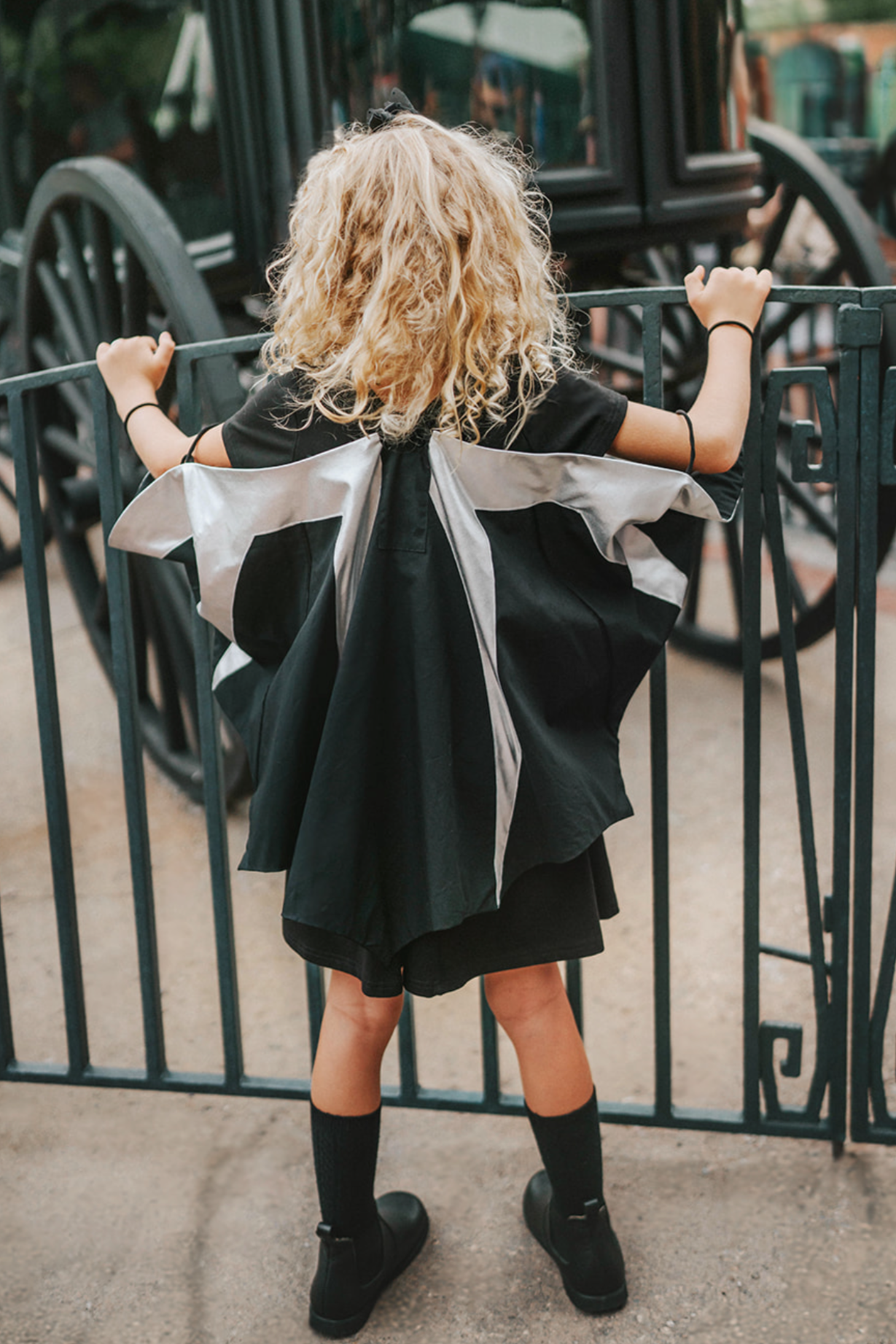 A young girl wears a solid black twirl dress.  There are removable bat wings fastened to the back.  They have metallic silver around the top and coming down to the bottom in long lines. There is elastic on the wings to put arms in to so the wings can be held out in a cape style motion. Perfect for spooky season.