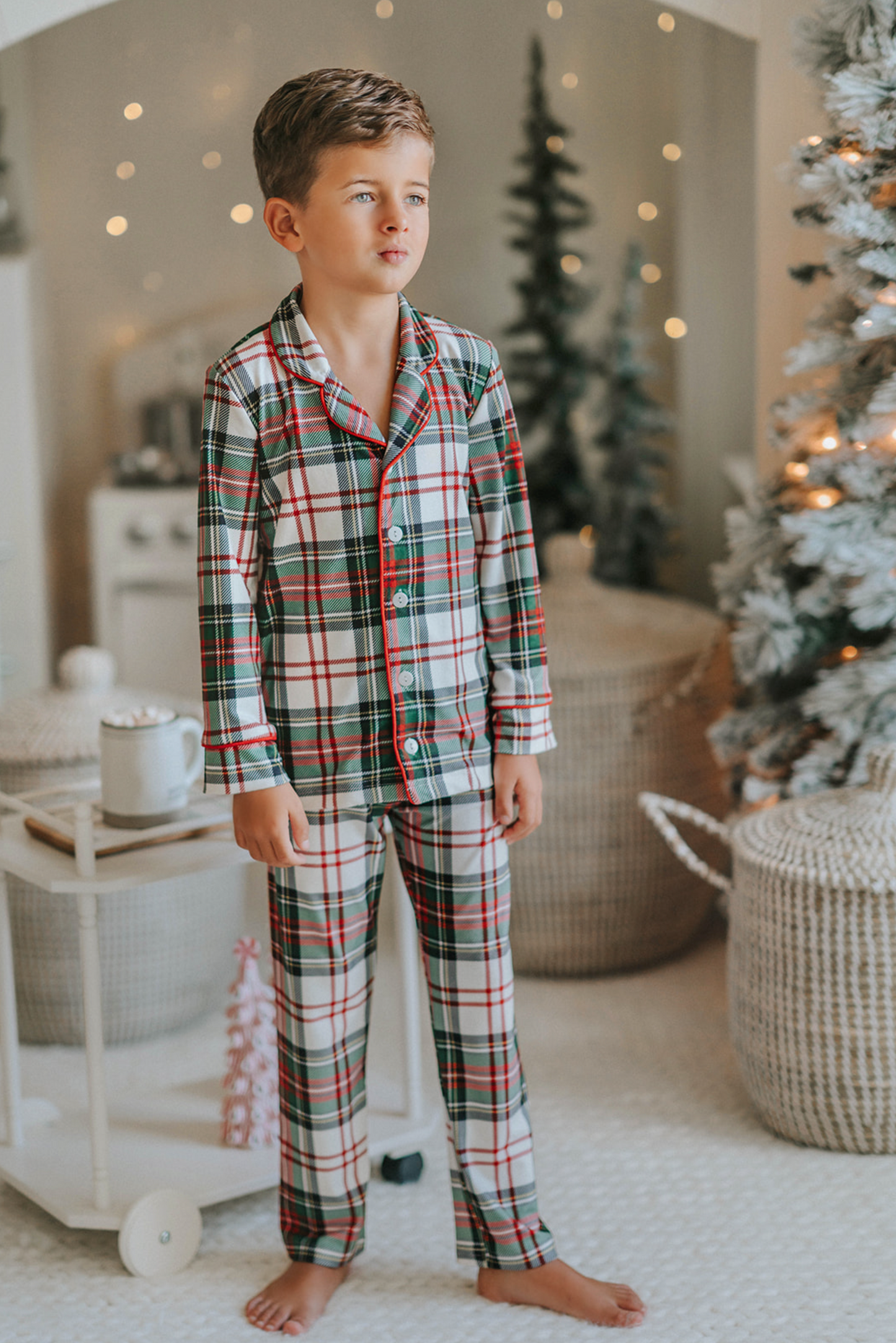 A young boy stands in a festively decorated room wearing Unisex Pj's in White Plaid. The classic 2 piece pajama set is buttery soft fabric.  The white background is accented by green, red and black plaid. There is red piping trim on the top. 