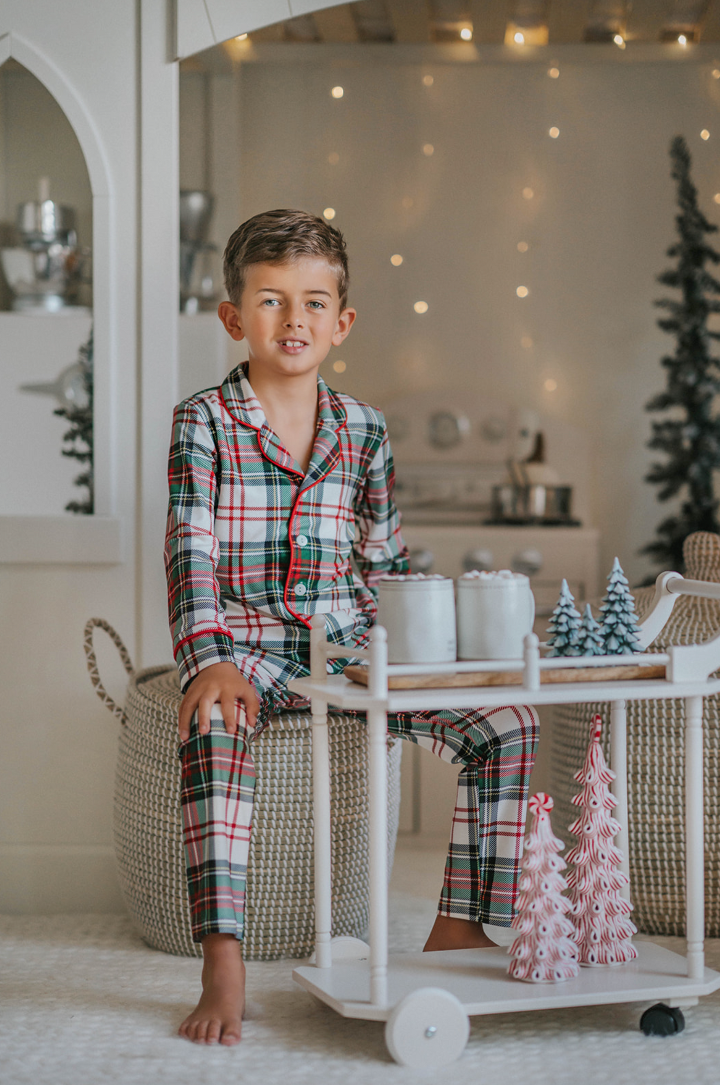 A young boy stands in a festively decorated room wearing Unisex Pj's in White Plaid. The classic 2 piece pajama set is buttery soft fabric.  The white background is accented by green, red and black plaid. There is red piping trim on the top. 