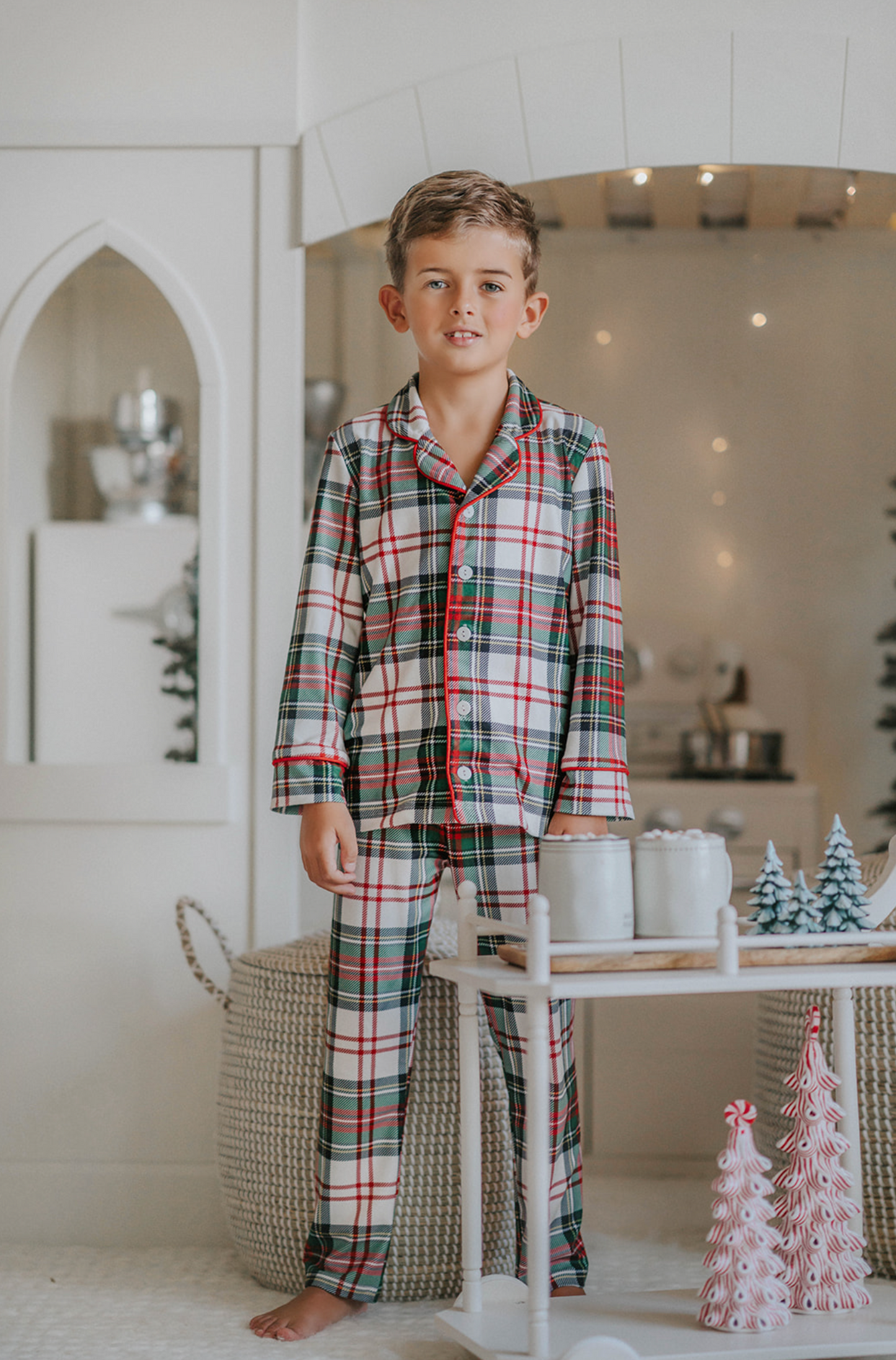 A young boy stands in a festively decorated room wearing Unisex Pj's in White Plaid. The classic 2 piece pajama set is buttery soft fabric.  The white background is accented by green, red and black plaid. There is red piping trim on the top. 