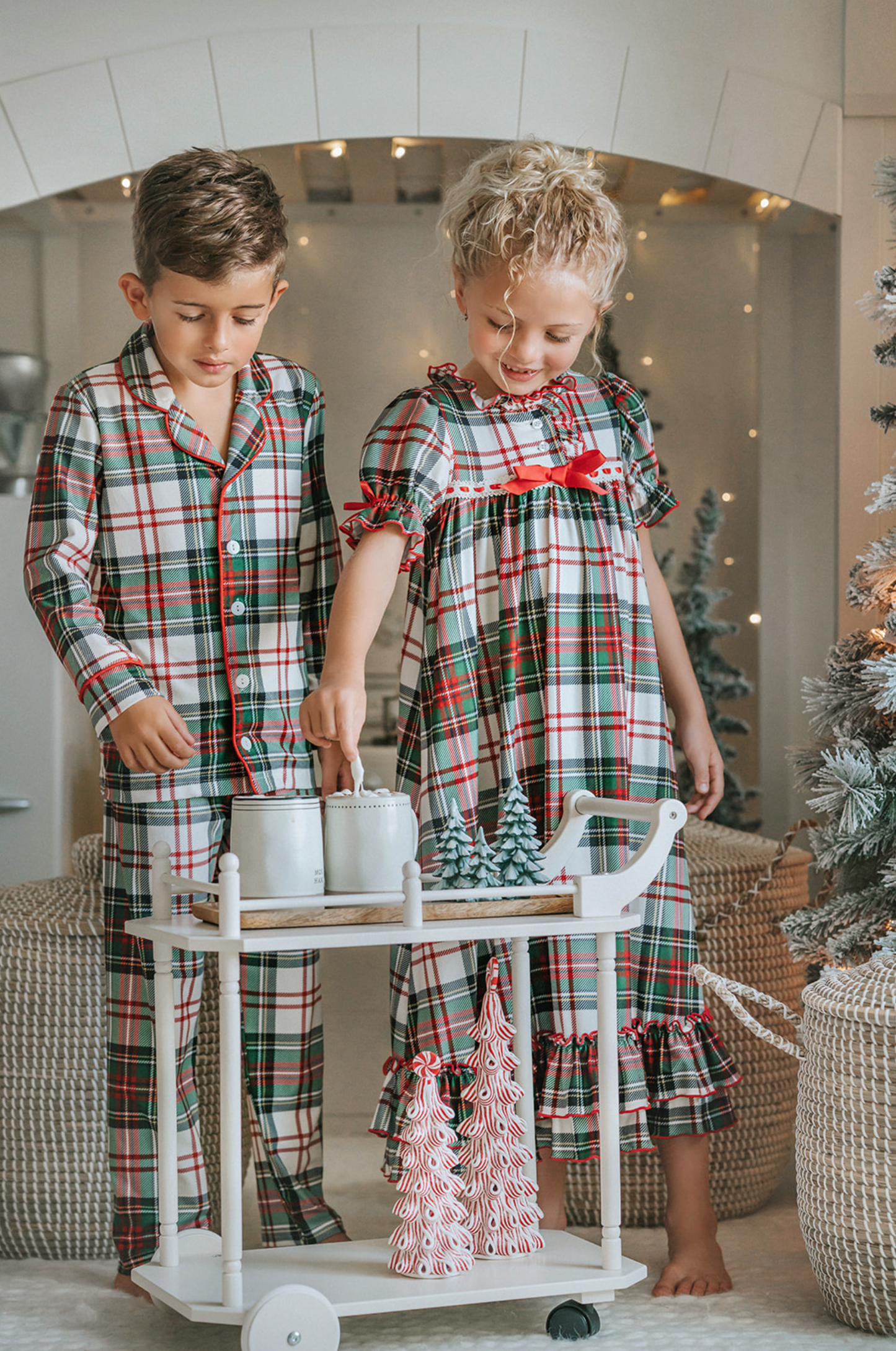 A young boy stands in a festively decorated room wearing Unisex Pj's in White Plaid. The classic 2 piece pajama set is buttery soft fabric.  The white background is accented by green, red and black plaid. There is red piping trim on the top. 