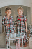 A young boy stands in a festively decorated room wearing Unisex Pj's in White Plaid. The classic 2 piece pajama set is buttery soft fabric.  The white background is accented by green, red and black plaid. There is red piping trim on the top. 