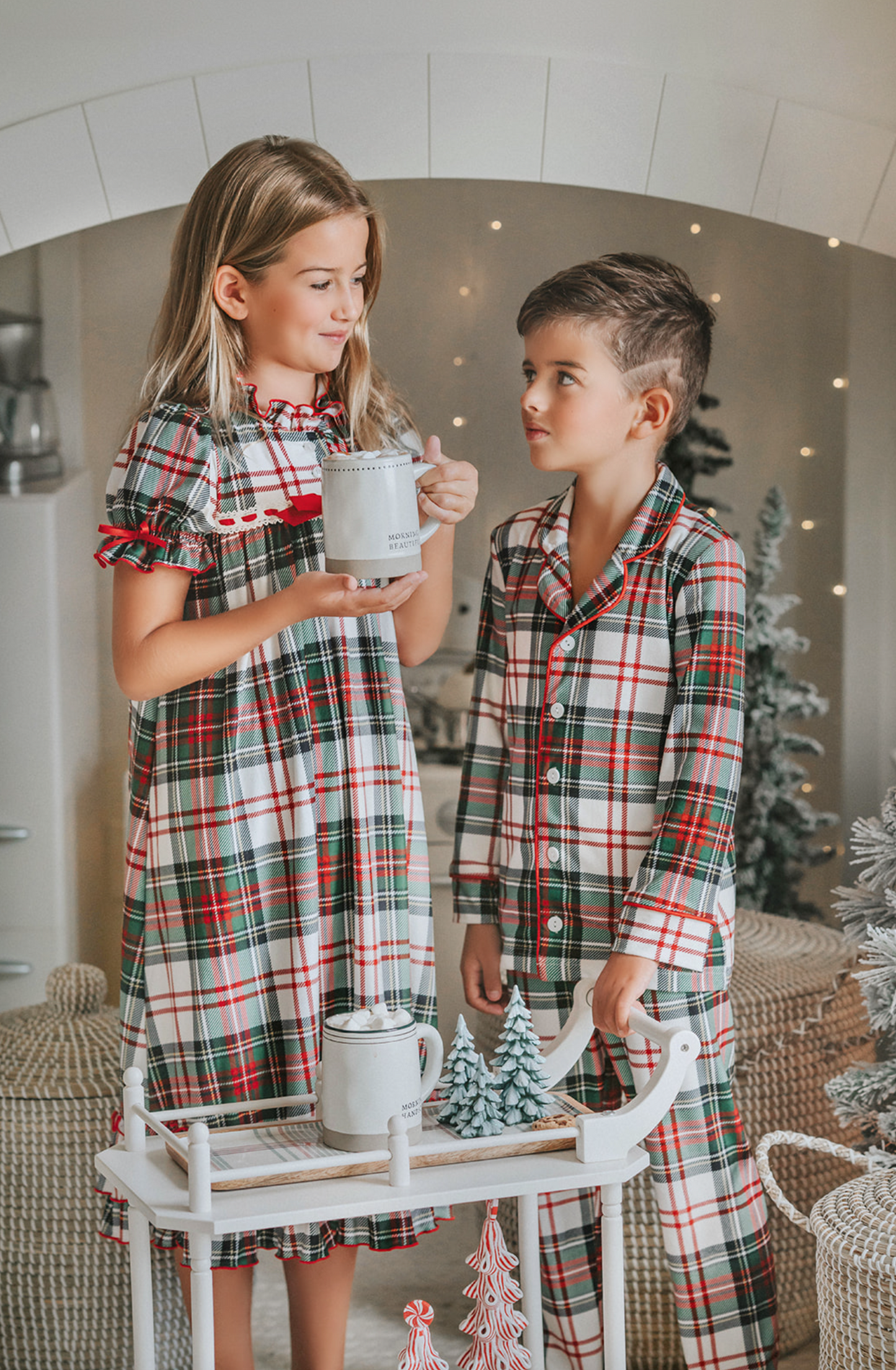 A young boy stands in a festively decorated room wearing Unisex Pj's in White Plaid. The classic 2 piece pajama set is buttery soft fabric.  The white background is accented by green, red and black plaid. There is red piping trim on the top. 